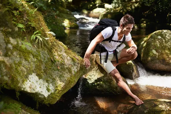 Man, hiking and river in tropical jungle with smile for adventure, journey and rocks with backpack. Person, outdoor and bag by water, earth and trees in rainforest on holiday with freedom in Colombia.