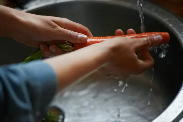 Hands, carrots and washing vegetables or healthy food in sink, preparing meal and kitchen while ready to cook. Person, rinsing fresh produce in basin at home for diet or organic recipe for wellness.