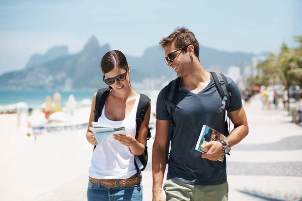 Couple, tourist and beach with map for guide on holiday, tour and travel for summer in Rio de Janeiro. Relationship, vacation and happy in seaside for trip in adventure, together and smile for break.