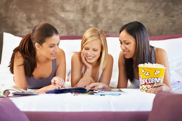 stock image Smile, friends and women reading magazine in bedroom, food and eating popcorn snack together at party. Happy, girls and group with journal in home for conversation or bonding in bed to relax in house.