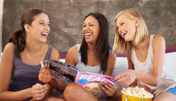 stock image Friends, laughing and magazine on bed with happiness, girls night and entertainment in home with popcorn. Women, paper and excited with snack in house for bonding, conversation and activity together.