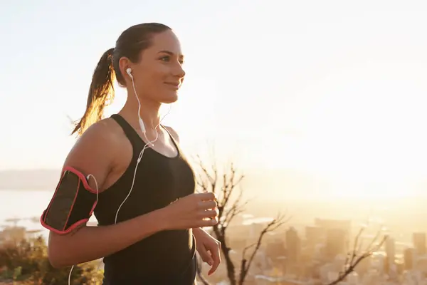 Woman, running and sunset with earphones on mountain for workout, exercise or cardio training. Active female person or runner listening to music, podcast or audio for motivation, health and wellness.