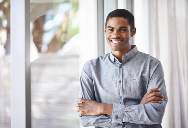 Happy black man, portrait and window with arms crossed for career ambition, job or creative startup at office. African male person or employee smile in confidence for expertise or positive mindset.