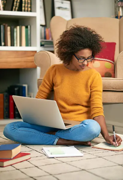 Laptop, books and African woman in library, floor and writing notes for education. Study, learning and female student with natural afro hair, assignment or technology for knowledge and reading.