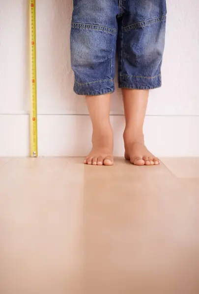 Child, wall and kid with tape measure for height, tracking growth or progress with feet in house. Legs, toddler and little person with development for size, length and stand on wooden floor at home.