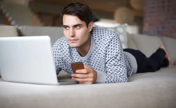 Man, laptop and smartphone at home on sofa for remote work as freelance, journalist and online research for newspaper. Male person, living room and focus on internet with information for article