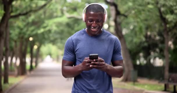 快乐的黑人男人在公园里带着耳机 音乐或聊天 森林中的智能手机 搜索引擎和男性跑步者 其应用程序可用于运动跟踪 进展或运动目标 — 图库视频影像
