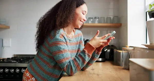 Woman, kitchen and typing on phone or browsing social media at home, happy and relaxing on weekend. Female person, communication and mobile application for online conversation, humor and laughing.