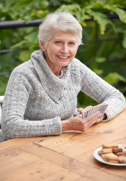 Elderly woman, cookies and portrait with cards at table for relaxation, old age and retirement. Senior person or grandmother and smiling with wrinkles for poker game, gamble and hobby for leisure.