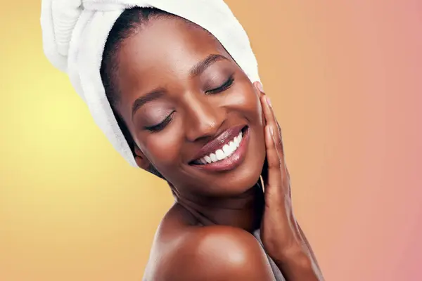 Beauty, hair towel or happy black woman in studio for skincare, wellness and makeup on orange background. Dermatology, cleaning or hands on face of African female model with cosmetic, shine or glow.