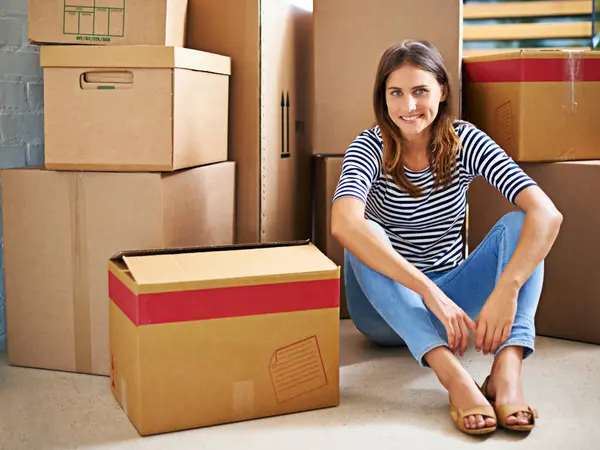 Confident, boxes and portrait of woman in new home for moving to estate, property or apartment. Happy, smile and young female person with cardboard package for equipment and products in modern house