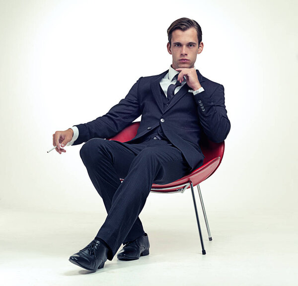 Suit, smoking and man portrait in chair, studio and serious in formal fashion by white background. Young model, face and elegant clothes with cigarette in armchair and confident in tuxedo by party.