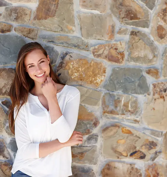Woman, portrait and smile by stone wall in home with confidence, relax or casual outfit on mockup space. Person, face and happy for holiday, vacation or peaceful getaway with shirt, good mood or calm.