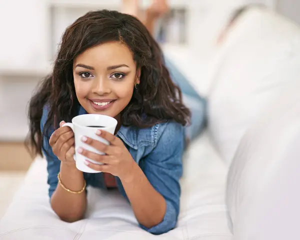 Portrait, coffee and relax with woman on sofa in living room of home for morning or weekend time off. Smile, beverage or drink with happy young person lying on couch in apartment for wellness.