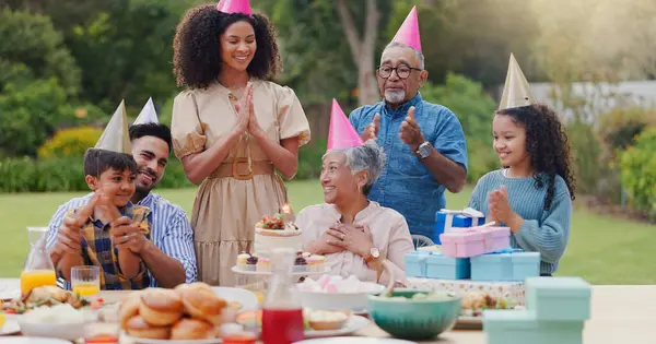 Family, clapping hands and happy with hat at birthday party for celebration, surprise or sparkler in garden of home. Grandparents, parents and kids with applause for gathering and event in backyard.