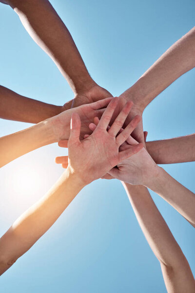 People, team and hands together below for unity, collaboration or synergy on a blue sky background. Closeup or low angle of group or community piling or stacking in solidarity for join or mission.