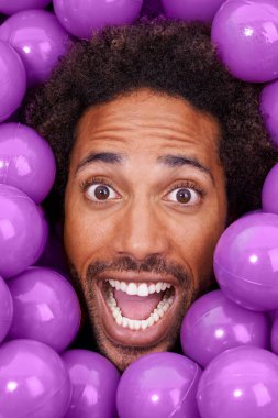 Ball pit, excited and portrait of black man with plastic toys for fun, playful and happy on background. Surprise, facial expression and face of person with purple balls, decoration and objects. clipart