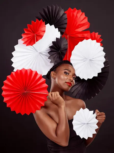 Black woman, origami fans and portrait in studio for beauty with makeup isolated on dark background. Cosmetics, pride and paper art for culture with tradition, skincare and confidence with elegance.