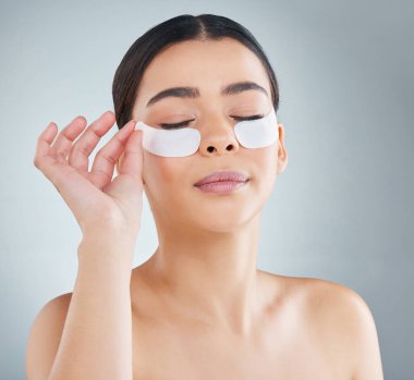 Woman, eye patch and hand in studio for skincare, dermatology and collagen or revitalise product for dark circles. Grey background, self care and cosmetic or facial beauty, antiaging and treatment. clipart