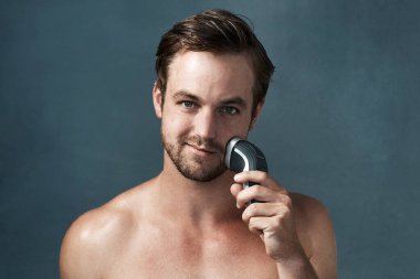 Stüdyoda tıraş makinesi, elektrikli ya da sakallı ya da cilt bakımı ya da kozmetik ürünleri için saç kesen bir adamın portresi. Yüzü, gülüşü ya da kendine güveni olan mavi arka plandaki kendine güvenen erkek güzelliği, yüz bakımı ya da sağlığı için.
