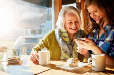 Üst düzey kadın, kızı sosyal medya anıları için kafede akıllı telefonuyla araştırma yapıyor ve gülümsüyor. Eğlenceli, postacı ve dinlendirici kahve, kek ve güneşli emeklilik ile heyecanlı