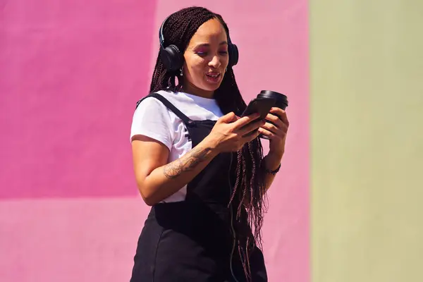 Stock image Woman, phone and outdoor for podcast and coffee, streaming music and subscription by wall background. Female person, latte and listening to podcast or online for social media, song and travel in city.