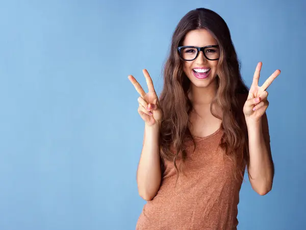 stock image Portrait, hands and happy woman with peace sign in studio for excitement, good mood or expression on blue background. V, emoji and model with glasses for gesture, positive attitude or smile mockup.