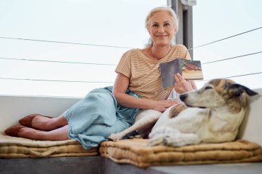 Yaşlı kadın, öğrenim, bilgi ya da balkonda birlikte rahat etmek için kanepede köpekle birlikte kitap ve portre okuyor. Hayvan, hikaye ve evcil hayvanı hobi, edebiyat ya da evde dinlenmek için evcil hayvanı olan yaşlı kişi..