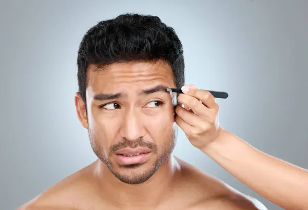 stock image Eyebrow, pain and man with tweezers in studio for hair removal, hygiene and facial treatment. Skincare, beauty and person anxious with tool for epilation, self care and grooming on white background.