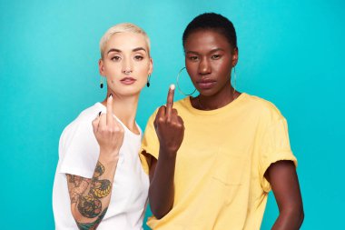 Women, portrait and middle finger with rude gesture in relationship, together and bonding in blue background. Female people, gay pride and serious for empowerment, diversity or inclusion in studio. clipart