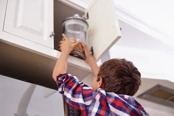 stock image Kitchen, cupboard and cookie jar with boy child reaching for treats for hunger in home from below. Biscuits, food or snack in glass with young kid in apartment, stealing baked goods to eat from back.