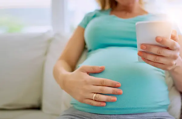 stock image Belly, hands and pregnant woman on sofa with cellphone for maternity research, information or blog. Pregnancy, relax and stomach of female person with phone for online prenatal care ebook at home