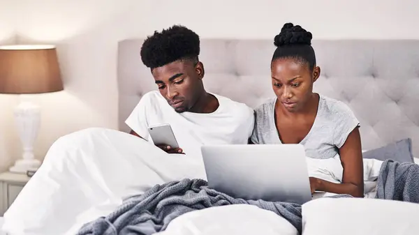 Stock image Black couple, laptop and bedroom with mobile phone, together in communication in morning. African people, wake up and tech for streaming, online activity and connection for movies or social media app.