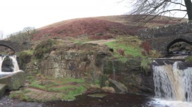 İngiltere 'nin Peak District Ulusal Parkı' ndaki Three Shires Head 'deki şelalelerin ve yük taşından köprülerin kırsal manzarasının solunda..