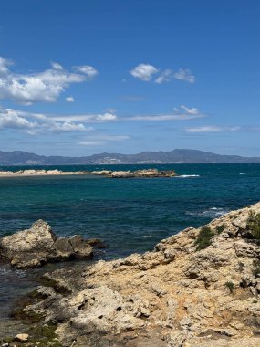 Arka planda mavi bir okyanus olan kayalık bir kıyı şeridi. Kayalar kıyı şeridi boyunca dağılmış ve su sakin. Sahne huzurlu ve sakin, okyanusla birlikte