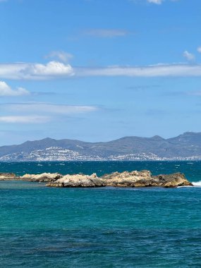 Arka planda mavi bir okyanus olan kayalık bir kıyı şeridi. Gökyüzü açık ve güneş parlıyor.