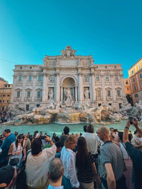 Büyük bir binanın önündeki çeşmenin etrafında büyük bir kalabalık toplandı. İnsanlar fotoğraf çekiyor ve manzaranın tadını çıkarıyor.
