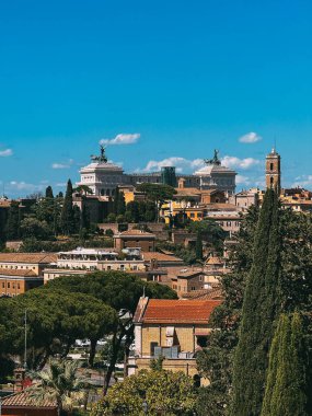Arkasında büyük bir bina olan bir şehir manzarası. Gökyüzü açık ve mavi. Ağaçlar yeşil, evler kahverengi.