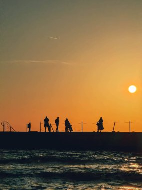 Gün batımında bir grup insan iskelede duruyor. Gökyüzü turuncu ve güneş batıyor.