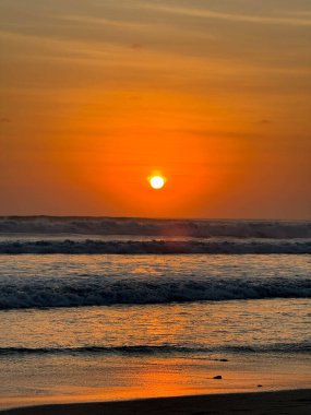 Güneş okyanusun üzerinde batıyor, suyun üzerine sıcak bir parıltı saçıyor. Dalgalar yumuşaktır ve gökyüzü turuncu ve pembe renklerin bir karışımıdır. Huzurlu ve huzurlu bir sahne.