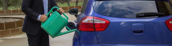 Image of an elegant man pouring water into the car tank with a sprinkler. Reference to the high cost of fuel and the transition to hydrogen cars. Horizontal banner