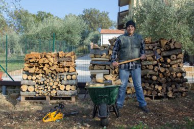 Elinde baltayla gülen bir oduncunun el arabasına odun yükledikten sonraki görüntüsü. Kış için evinizi ısıtmak için stok yapın.