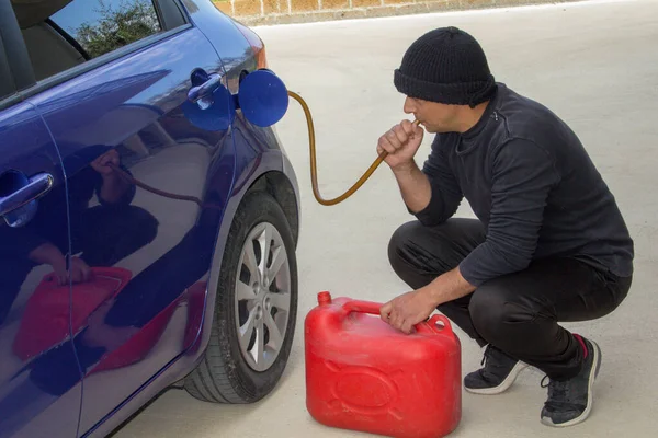 Image of a thief dressed in black who sucks petrol from a car tank with a rubber hose. Reference to the increase in fuel theft due to excessive cost.