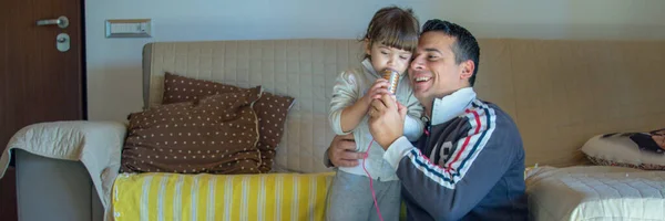 Image of a smiling dad hugging his daughter as they play house and sing karaoke. Horizontal banner