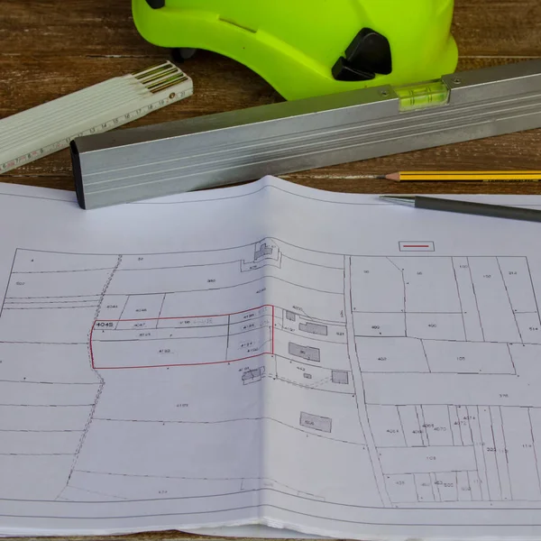 Image of a work table where plans for a building site are placed, a work helmet, a level, tape measure and pencils. Construction engineer job