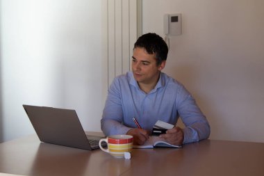 Image of a smiling man behind a desk typing on an agent and holding credit cards after shopping online from his computer.