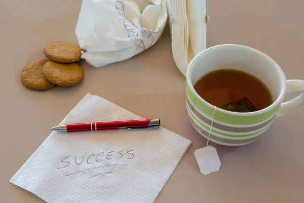 Image of a napkin with success written on it, and cookies in the background, a cup of tea, a pen and more napkins. Reference to ambitions and ideas during breakfast.