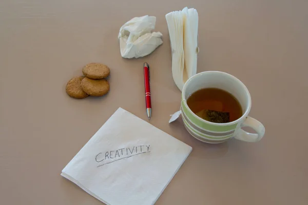 Image of a napkin with creativity written and in the background, a cup of tea, a pen, biscuits and other napkin scribbles. Reference to ambitions and ideas during breakfast.