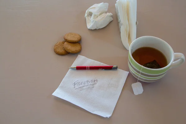 Image of a napkin with freedom written and in the background, a cup of tea, a pen, biscuits and other napkin scribbles. Reference to ambitions and ideas during breakfast.