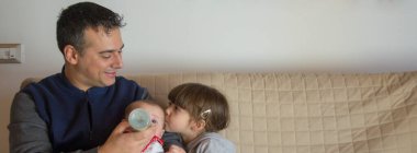 Evdeki kanepede oturan bir babanın yeni doğmuş oğlunu kucağında tutarken Bineron ve ablasının ona sarılıp öptüğü görüntüsü. Kardeş sevgisi ve sevgi dolu bir aile. Yatay Pankart 